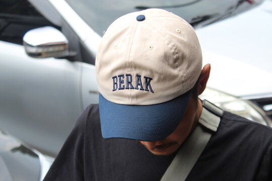 Close-up of a person wearing a casual cap featuring a humorous Indonesian slang word "BERAK". The word is a crude slang for defecation in Indonesia