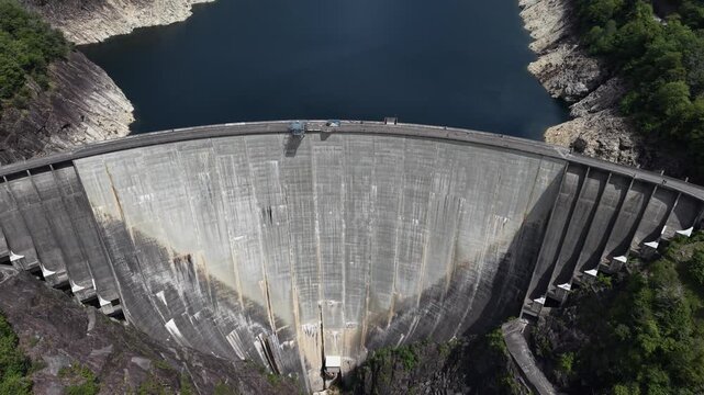 Aerial view of the impressive concrete arch of the Verzasca Dam surrounded by steep rocky cliffs and dark reservoir water in Gordola, Ticino, Switzerland.