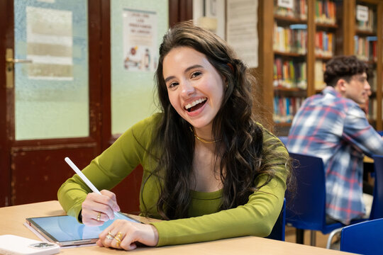 Smiling young woman learning and studying with a digital tablet and stylus in a high school library