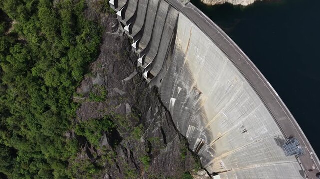 Aerial view of the massive Verzasca Dam surrounded by steep rocky cliffs and lush green forests in Gordola, Ticino, Switzerland.
