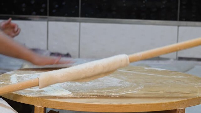 An authentic culinary culture video showcasing the skilled hands of a woman rolling out dough using traditional methods and the process of making yufka on a wooden floor mat dusted with flour.
