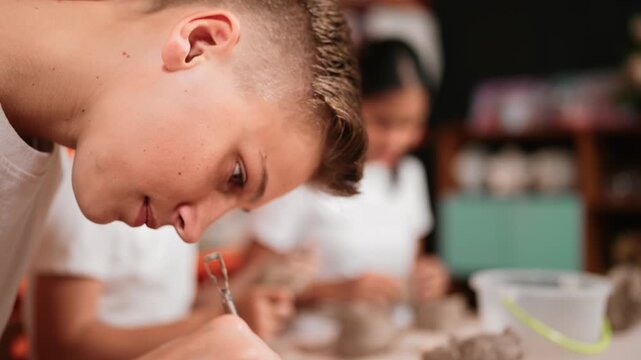 Closeup of high school boy hand using carving tool sculpt cup of clay. Diverse children doing activity while playing or modeling piece of clay in pottery workshop. Blurring background. Edification