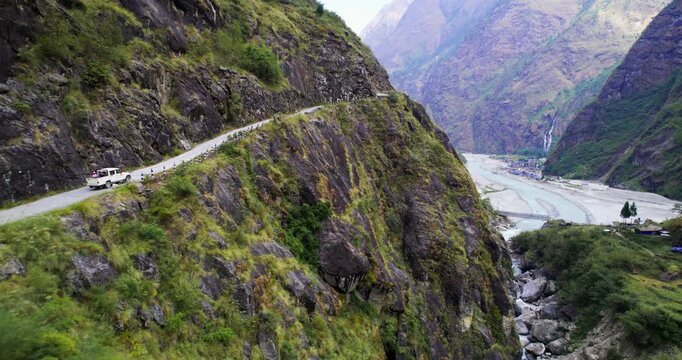 Aerial view of a white 4x4 driving along a cliffside road above a turquoise river winding through a lush green Himalayan valley in Nepal.
