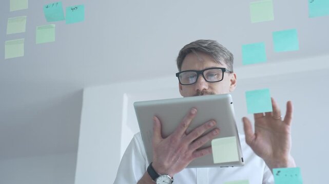 Adult businessman in glasses using a tablet computer while brainstorming and organizing work on a transparent glass board with colorful sticky notes, focusing on strategy and planning