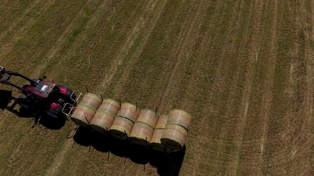 Tractor Harvesting Hay Bales in Field: An aerial view captures a tractor hauling a line of freshly baled hay across a sun-drenched field.