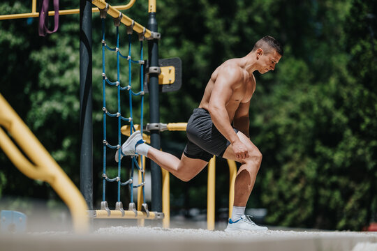 Shirtless man performs a split squat with his rear foot on a rope net in an outdoor park. Outdoor workout demonstrates strength, balance and leg training using park equipment.