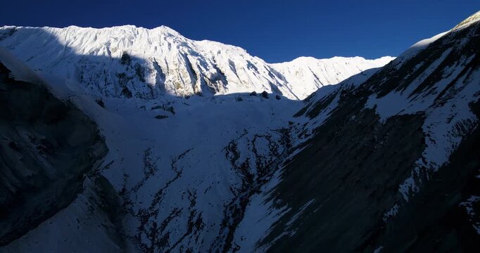 A dramatic aerial drone view captures the massive snow-covered ridges and steep faces of a Himalayan mountain pass under a deep blue sky in Nepal.