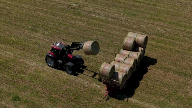 Tractor Moving Hay Bales in a Field: A tractor with a front loader lifts a round hay bale, moving it towards a trailer loaded with more bales in a vast, sunlit field.