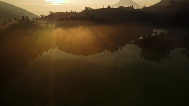 Aerial view of Dieng Plateau with thick golden mist reflecting on the calm lake surface surrounded by dark hills at sunrise in Kejajar, Central Java, Indonesia.
