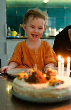 A 3 year old toddler boy is making a wish before blowing out the candles on his birthday cake. The DIY cake features a fun construction site decoration with toy trucks.