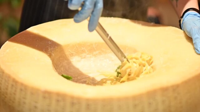 Chef mixing fresh pasta inside large hollowed parmesan cheese wheel in restaurant kitchen.
