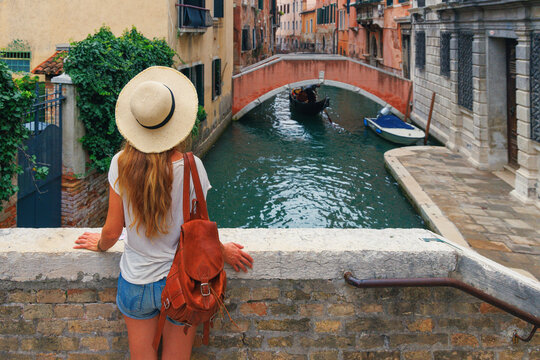 back view of a female tourist in straw hat with backpack leaning on a stone bridge looking at gondola in venice canal italy
