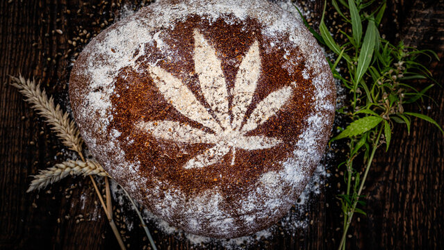 Freshly baked bread with a cannabis leaf design on the crust, placed on a wooden surface with herbs and wheat