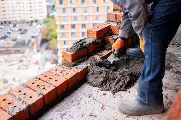 A construction worker wearing orange gloves and a grey uniform actively uses a hand trowel to apply mortar and lay red clay bricks on a high-rise urban building site. © Aigul Sabirova
