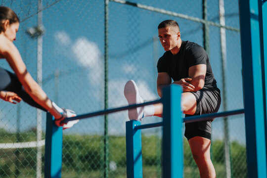 A man stretches his leg on parallel bars during an outdoor workout with a partner. A woman stretches on the bars nearby as they warm up for strength training.