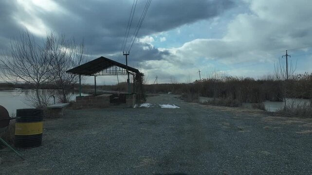 Camera moving forward on a gravel road next to a frozen lake on a cloudy winter day, with trees and electric poles on the side and mountains in the distance.

