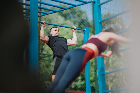 A muscular man performs a pull-up on a blue outdoor bar. A woman trains nearby on parallel bars in the same outdoor gym setting.