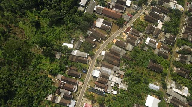 Top-down aerial view of a rural village road bordered by houses and lush green forests.
