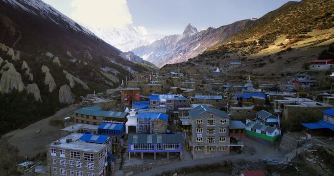 An aerial view of a Himalayan village with blue rooftops set in a valley beneath dramatic snow-capped peaks at golden hour in Nepal.