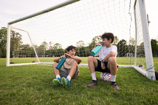 Two young boys drink from their water bottles while sitting on grassy field in front of soccer goal