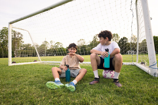 Two young boys drink from their water bottles while sitting on grassy field in front of soccer goal