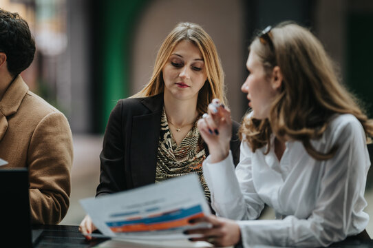 Two women colleagues review printed documents while discussing work. The pair consult charts and papers during a casual outdoor business meeting.