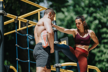 Fototapeta premium A fit woman stretches her leg against her male partner during an outdoor workout. They train together at a calisthenics park, practicing partner stretching and strength exercises.