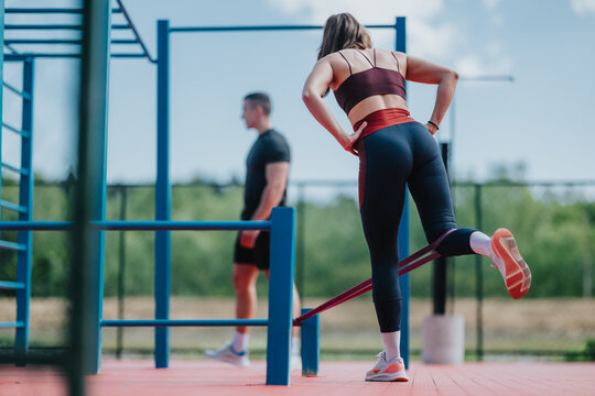 A woman performs a resistance-band leg exercise while a trainer watches in an outdoor gym. The athlete trains with bands on a fitness course for strength and balance.