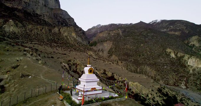 An aerial drone view captures a white Buddhist stupa with prayer flags in the village of Manang, surrounded by the dramatic dry mountains of Nepal's Annapurna region.
