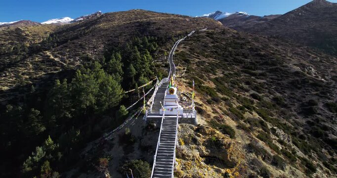 An aerial view of a white Buddhist stupa with colorful prayer flags and a stairway on a Himalayan ridge in Nepal.