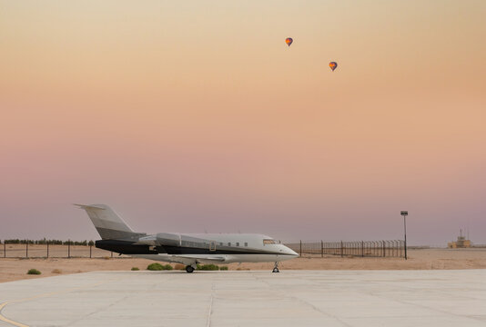 Private jet parked at desert airport runway with hot air balloons at sunset, luxury business travel scene