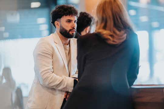 A young male professional speaks with a female colleague at a reception desk. The pair are engaged in a focused office conversation near the front desk.