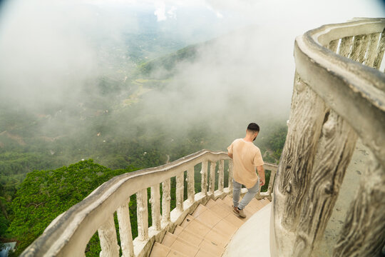 Sri Lanka - 22 February 2026: View of a man walking down a spiral staircase with white railings overlooking a lush green valley covered in thick fog.