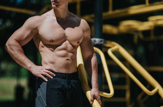 Shirtless muscular man with defined abs stands at outdoor gym equipment. He poses by yellow parallel bars during an outdoor workout session.