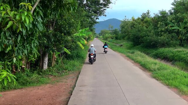 A dynamic drone follow shot capturing two motorbikes cruising along a peaceful, narrow paved road bordered by lush banana plantations and dense tropical greenery, set against a backdrop of distant rol