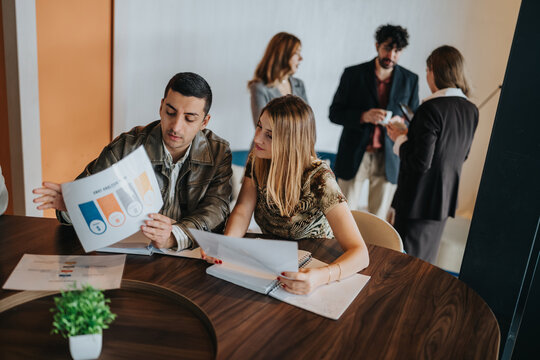 Two colleagues review business reports and charts at a round meeting table. Coworkers stand and converse in the background during a casual office meeting.