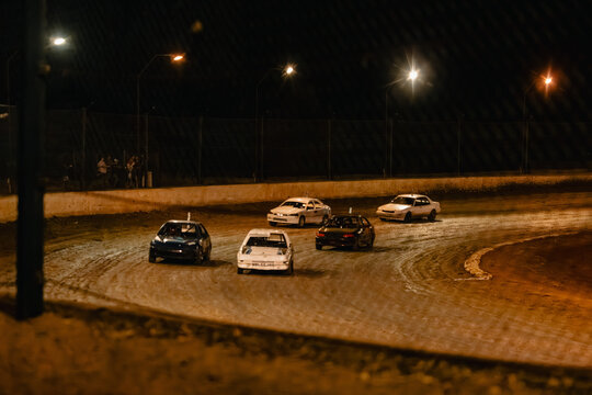 Stock cars race on a dirt track at night under bright stadium lights
