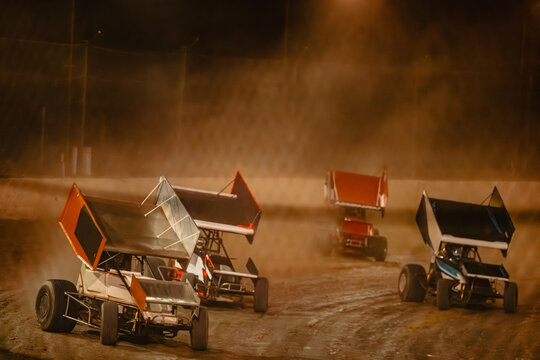 Sprint cars race on a dirt track at night kicking up dust under lights