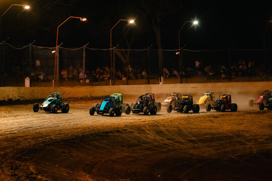 Sprint cars race on dirt track at night under bright stadium lights with spectators watching