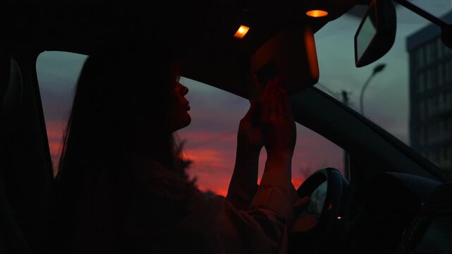 Woman car sunset silhouette adjusts sun visor mirror while sitting inside vehicle during vibrant red dusk in the city