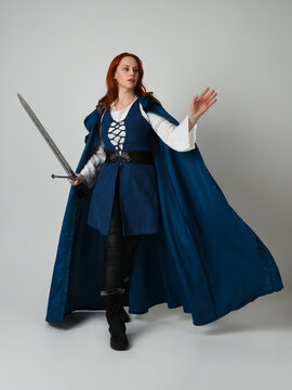 full length portrait of female model wearing medieval fantasy warrior historical costume with blue cloak. silhouette figure, holding sword fighting weapon on studio background.