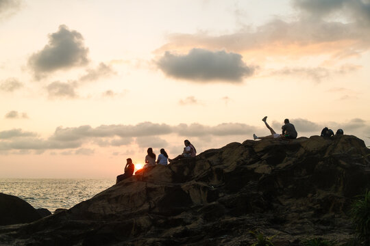 Sri Lanka - 25 January 2026: View of people sitting on a rocky cliff overlooking the ocean during a golden sunset with soft clouds.