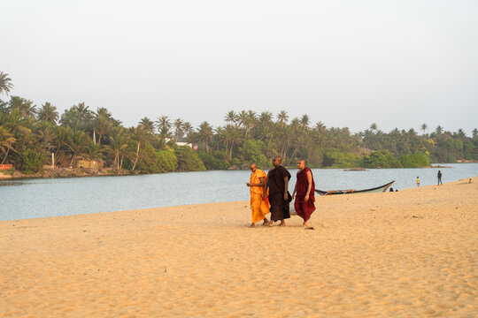 Sri Lanka - 23 January 2026: View of three Buddhist monks walking along a sandy beach by a tropical lagoon with palm trees and a boat in the background.