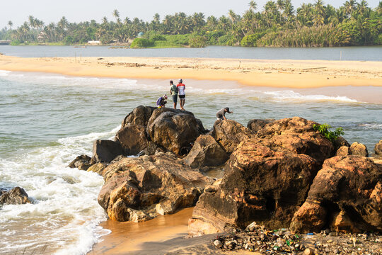 Sri Lanka - 21 January 2026: View of people standing on large coastal rocks as waves crash against the shore with a sandy beach and palm trees in the background.