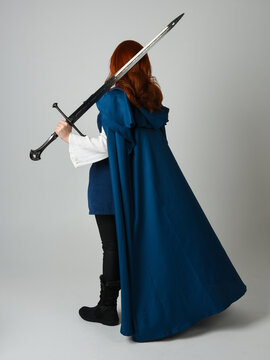 full length portrait of female red head model wearing medieval fantasy warrior historical costume with blue cloak. silhouette back view figure walking away and holding sword weapon, studio background