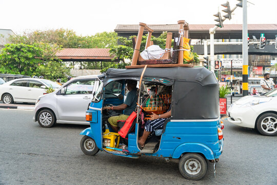 Sri Lanka - 15 January 2026: View of a blue tuk-tuk transporting a large wooden table on its roof through city traffic with cars and trees in the background.