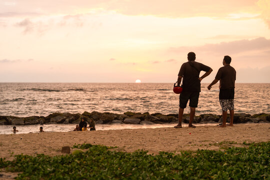 Sri Lanka - 14 January 2026: View of two men standing on a sandy beach watching the sunset over the Indian Ocean with people swimming near the shore.