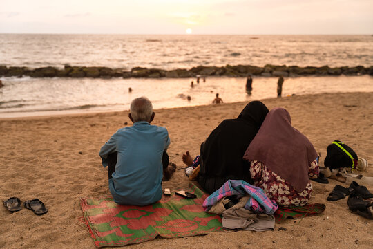 Sri Lanka - 14 January 2026: View of a man and two women sitting on the sand watching the sunset over the Indian Ocean with gentle waves and a soft sky.