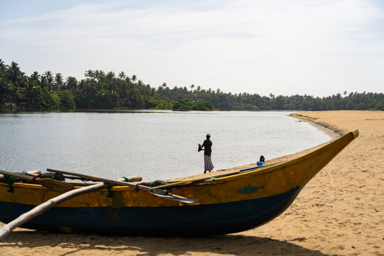 Sri Lanka - 09 January 2026: View of a traditional wooden outrigger boat on a sandy beach with a fisherman standing by the calm water and tropical palm trees.