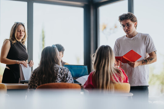 Teenage friends gather around a table sharing notes and a laptop while one presents from a notebook. A small group discussion and study session in a bright, modern space.